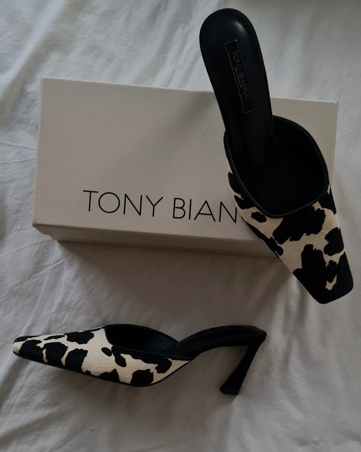 Cow print pointed-toe mule heels with stiletto heels next to a shoebox.