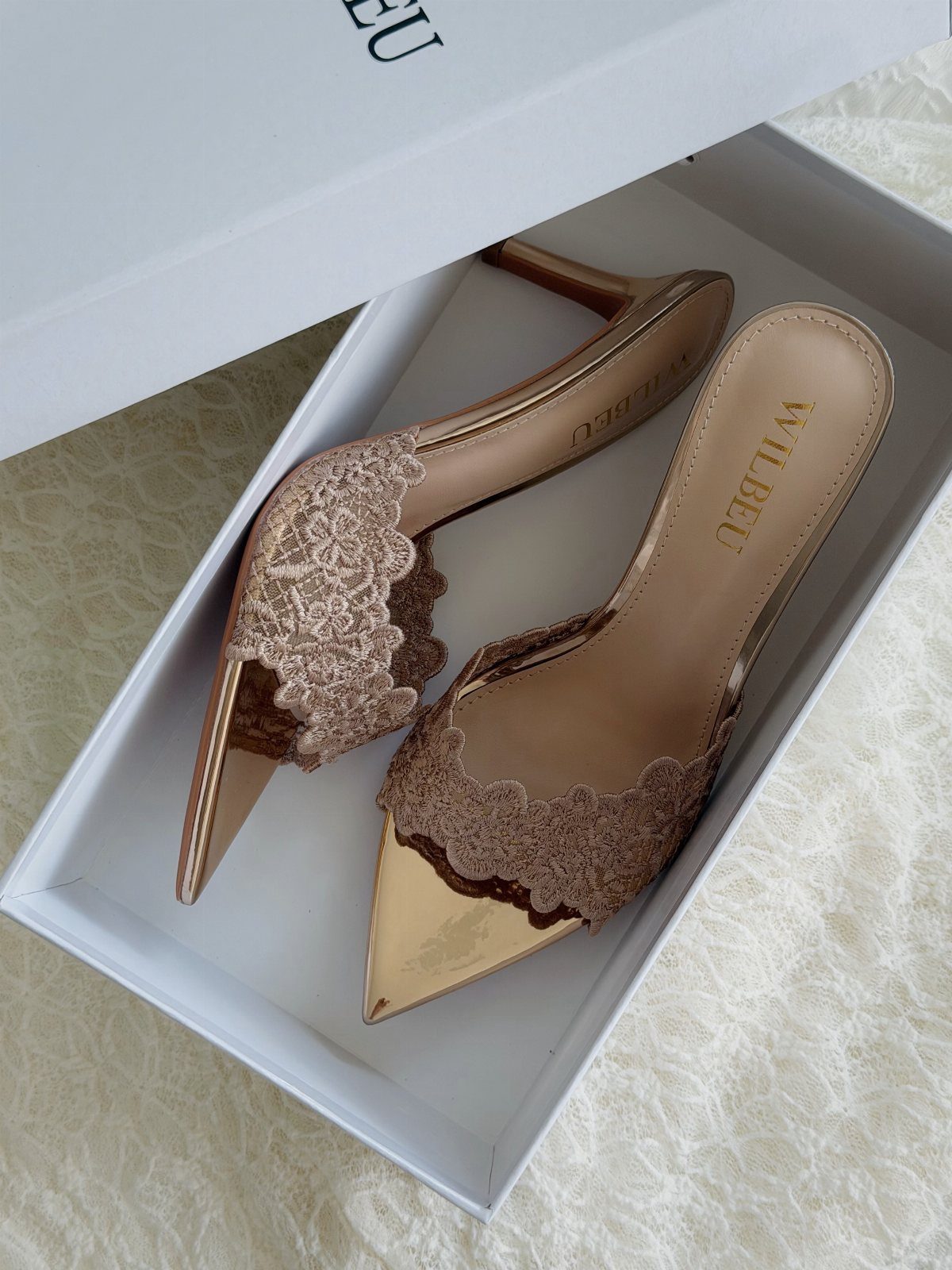 Gold pointed-toe mule heels with beige lace scalloped edges in a white box
