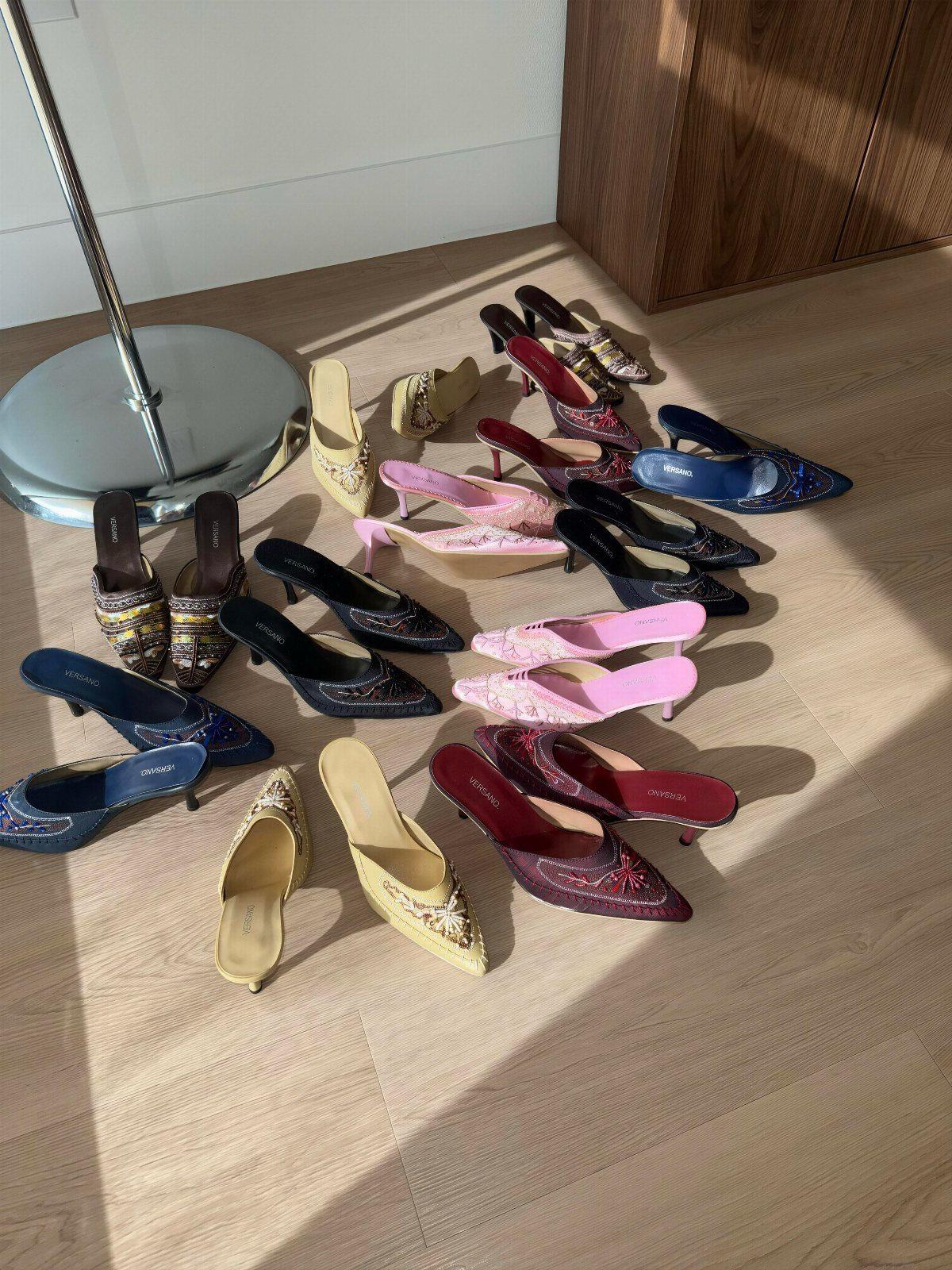 Assorted beaded heeled mules in various colors arranged on a light wooden floor.