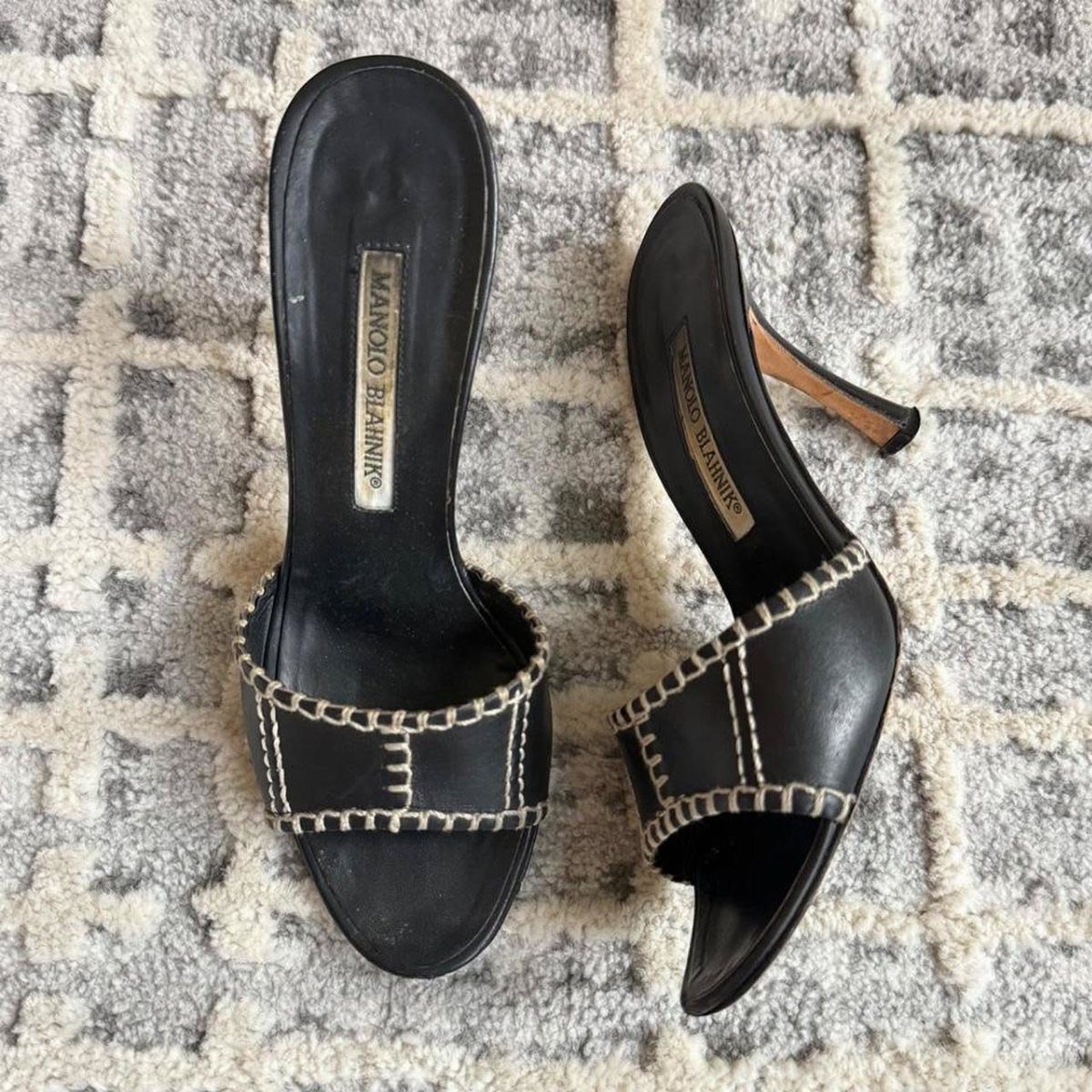 Black leather mule heels with white contrast stitching on a textured rug