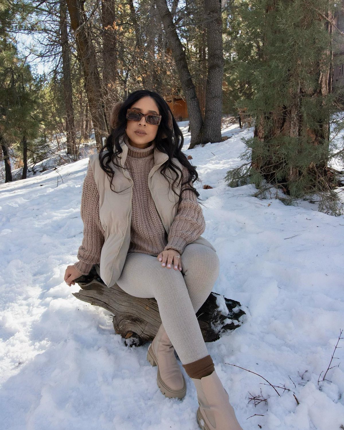 Woman in beige puffer vest and snow boots sitting on log in snow