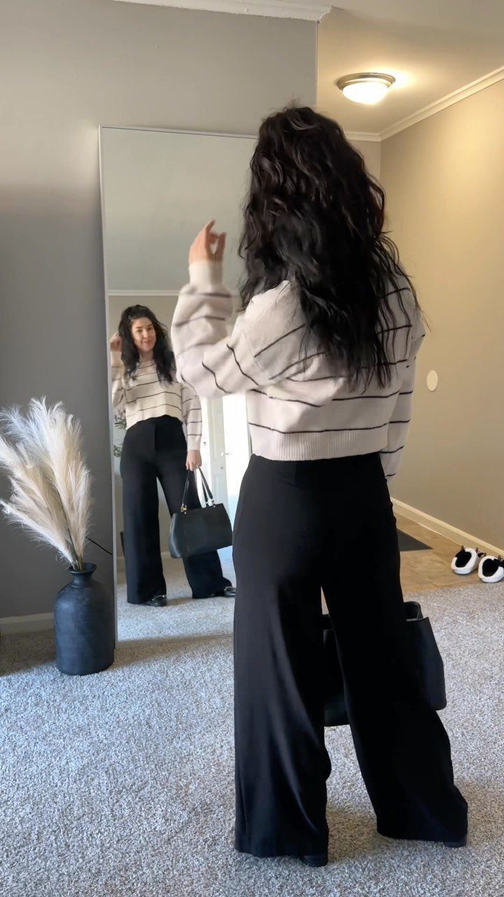 Woman wearing wide-leg black trousers and a striped sweater posing before a mirror.
