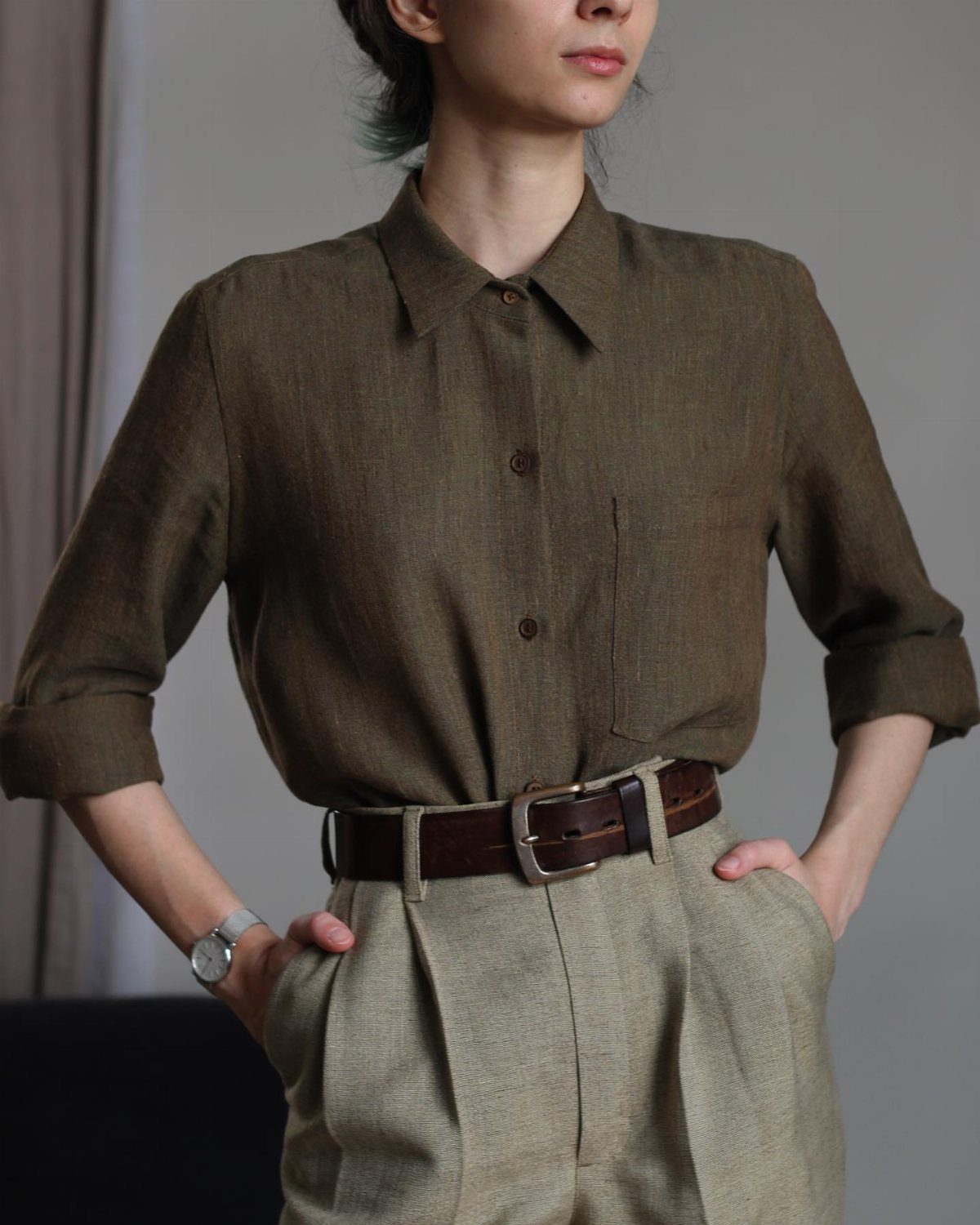 Woman wearing an olive green button-down shirt and beige pleated trousers with belt.
